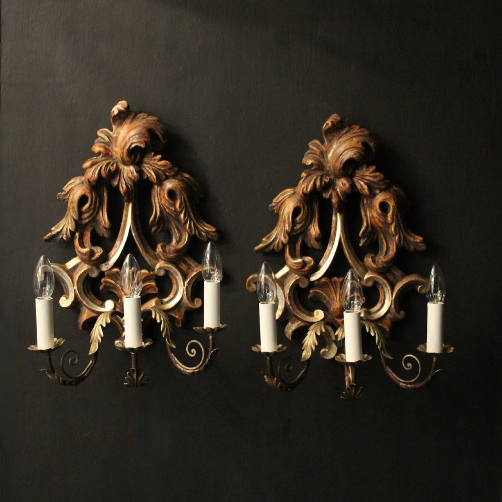 A pair of carved giltwood three-arm Florentine wall lights with ornate acanthus leaf scrollwork and candle sleeves.