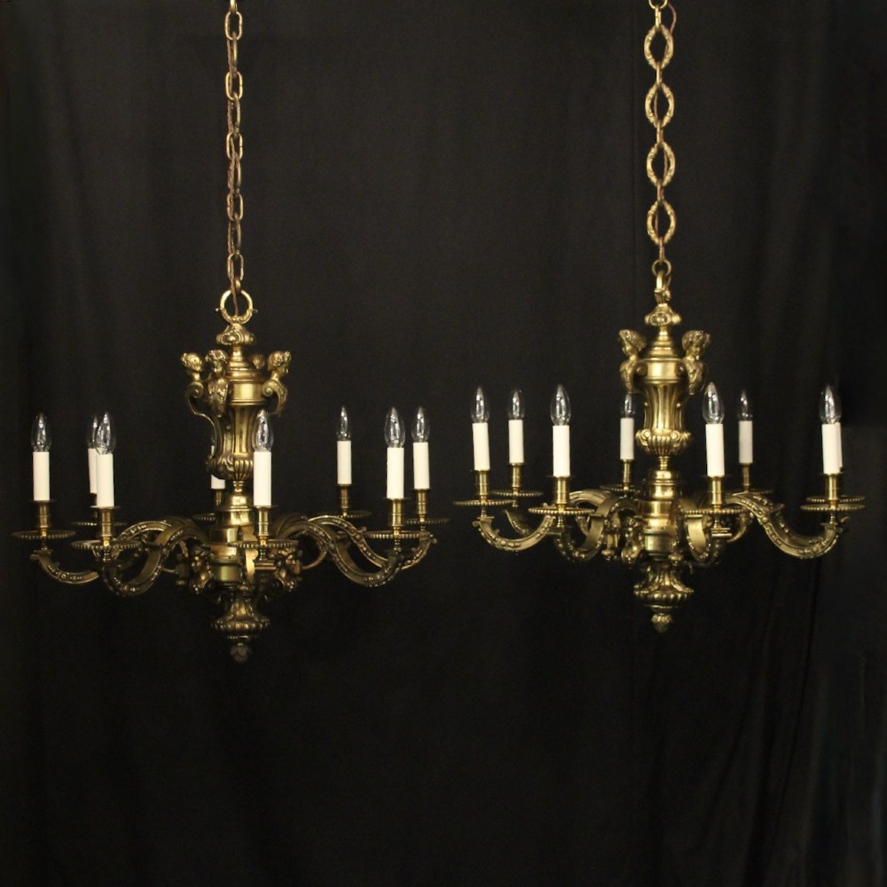A matching pair of cast bronze eight-light chandeliers featuring ornate scrolled arms, decorative stem castings, and faux candle sleeves.