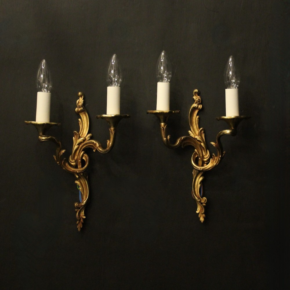 A pair of two-arm French Rococo style gilded bronze wall lights featuring acanthus leaf scrollwork and cream candle holders.