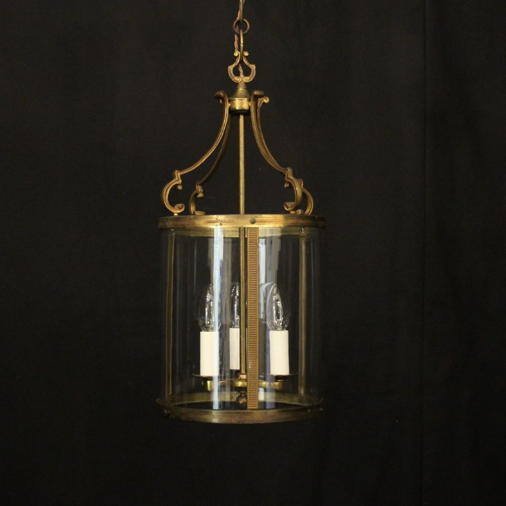 An antique French cylindrical hall lantern featuring a gilded brass frame, curved glass panels, and a three-light interior fitting suspended from a hook.