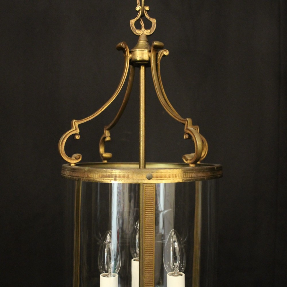 An antique French cylindrical hall lantern featuring a gilded brass frame, curved glass panels, and a three-light interior fitting suspended from a hook. View V