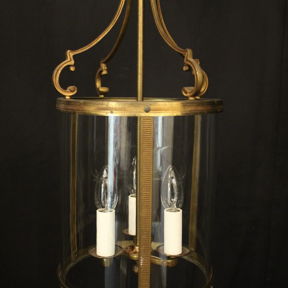 An antique French cylindrical hall lantern featuring a gilded brass frame, curved glass panels, and a three-light interior fitting suspended from a hook. View VI