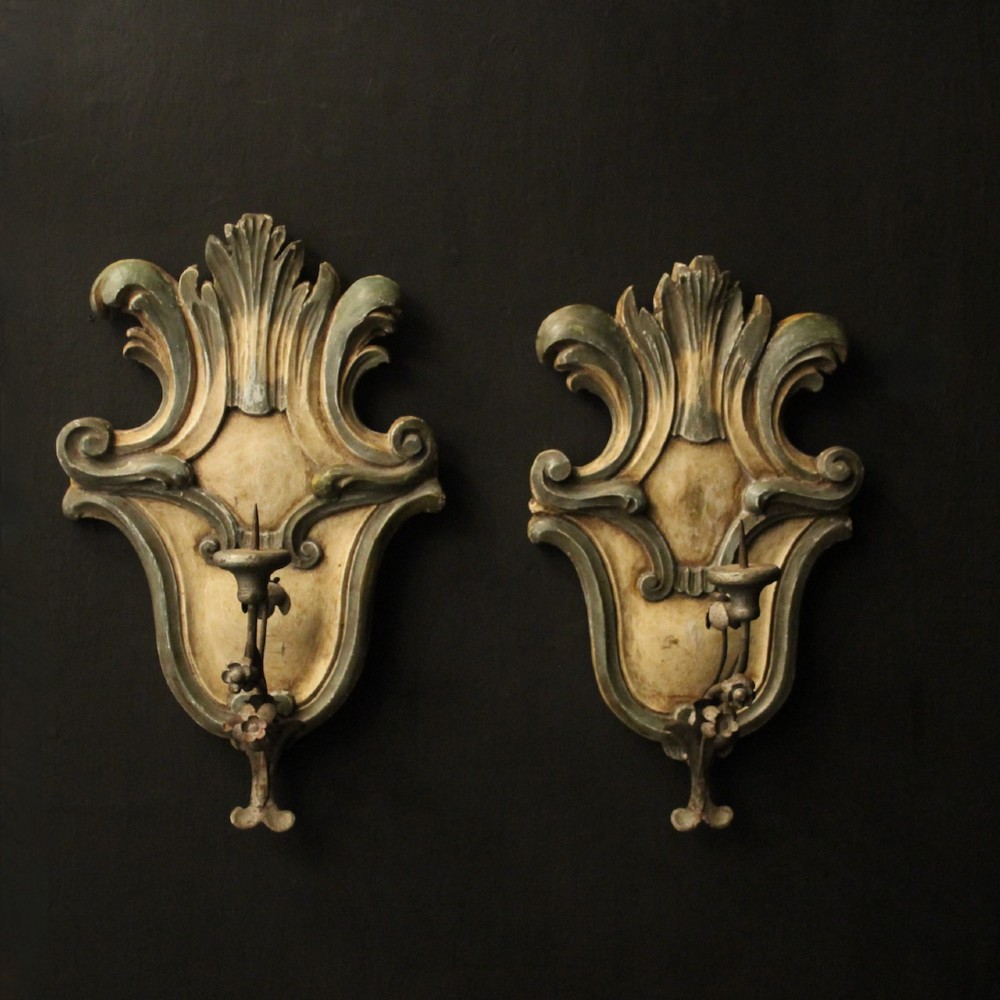 A pair of carved Italian Florentine wall sconces in distressed cream and sage green paint, featuring acanthus crests and single metal candle prickets.