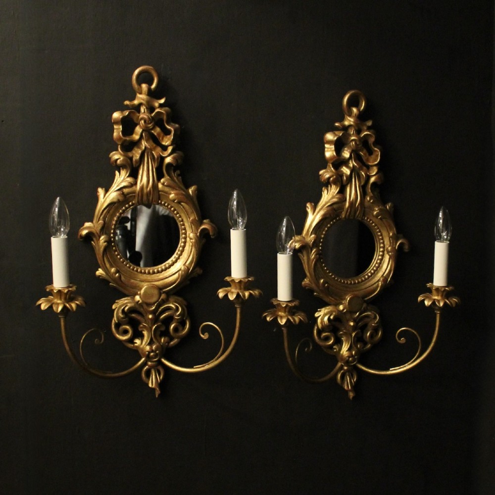 A pair of carved giltwood twin-arm girandole wall sconces featuring central circular mirrors, ornate scrollwork, and bow-tied cresting.