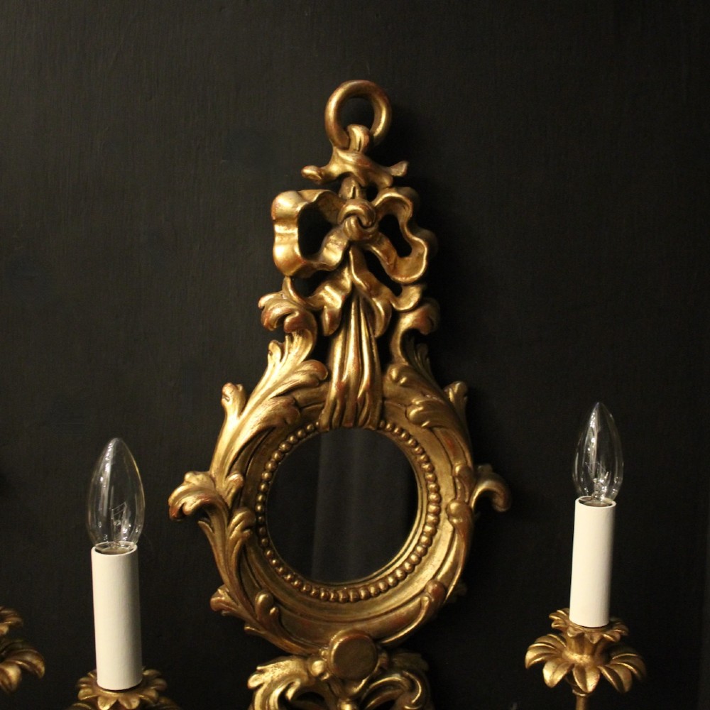 A pair of carved giltwood twin-arm girandole wall sconces featuring central circular mirrors, ornate scrollwork, and bow-tied cresting. View V