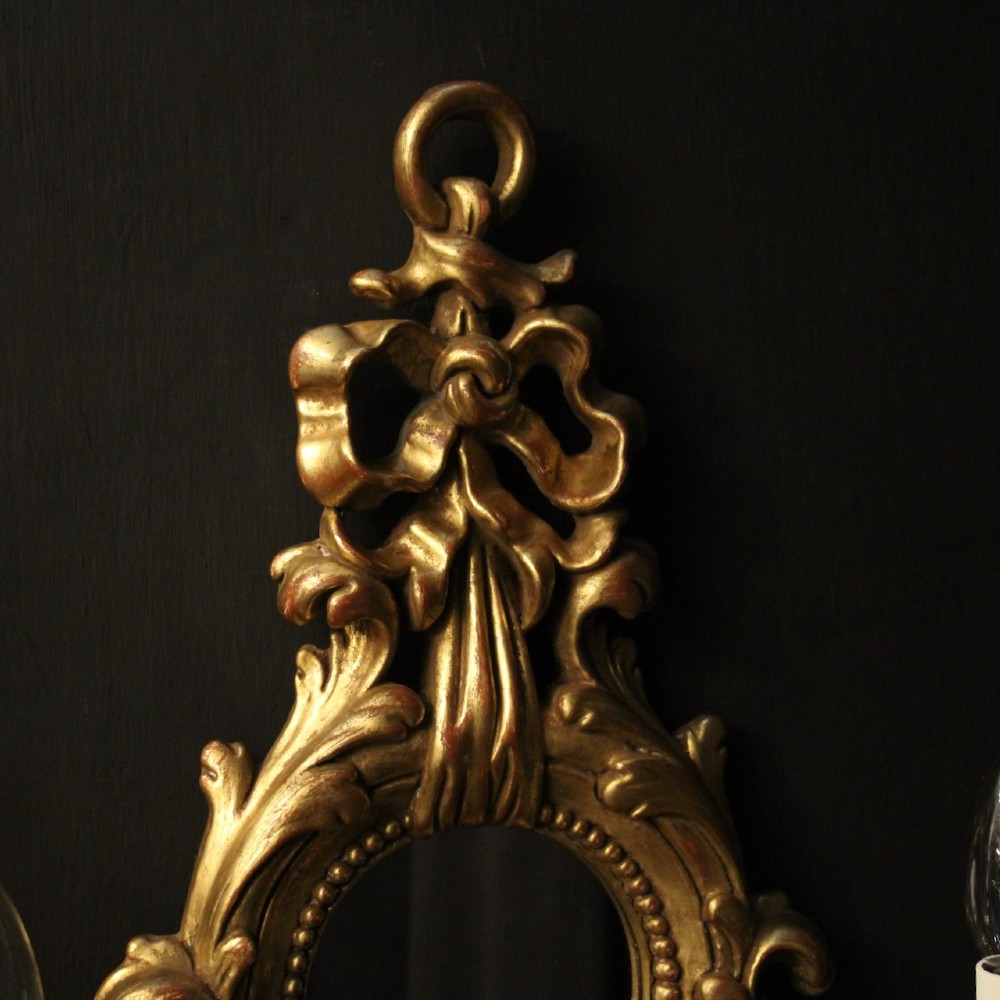 A pair of carved giltwood twin-arm girandole wall sconces featuring central circular mirrors, ornate scrollwork, and bow-tied cresting. View VI