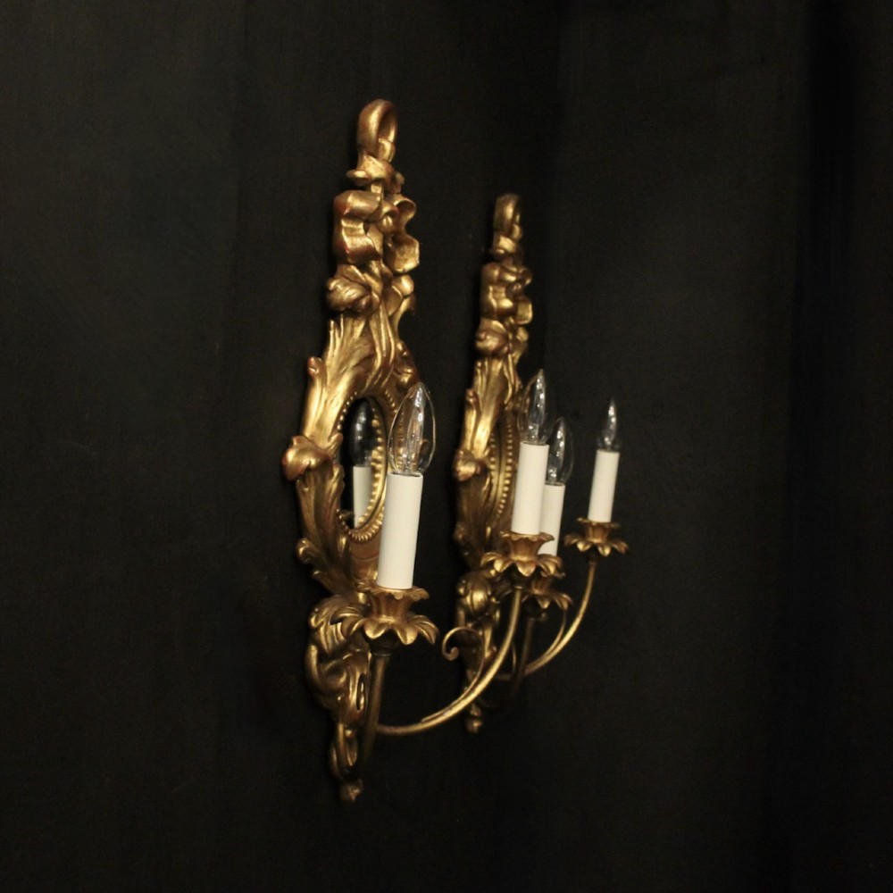A pair of carved giltwood twin-arm girandole wall sconces featuring central circular mirrors, ornate scrollwork, and bow-tied cresting. View X