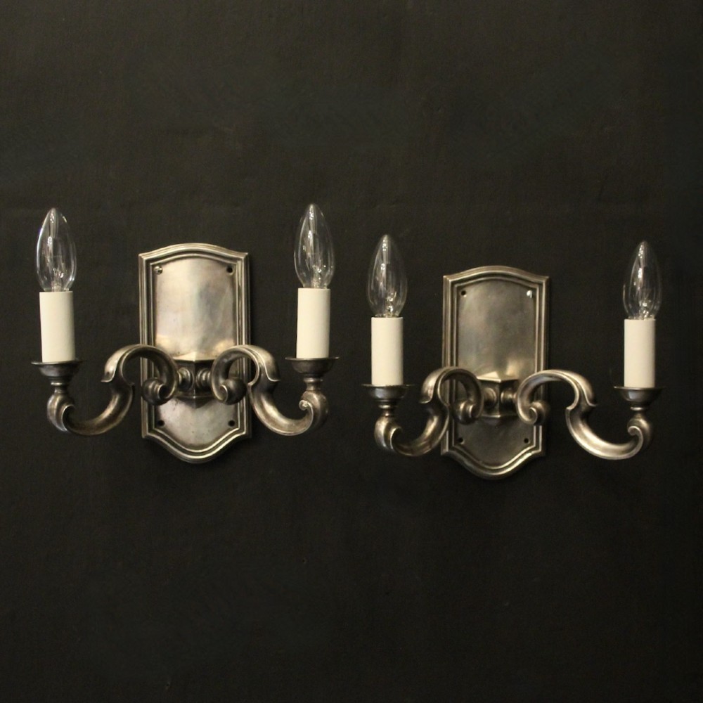 A pair of antique silver-plated twin-branch wall sconces featuring scrolling arms and moulded backplates against a dark background.