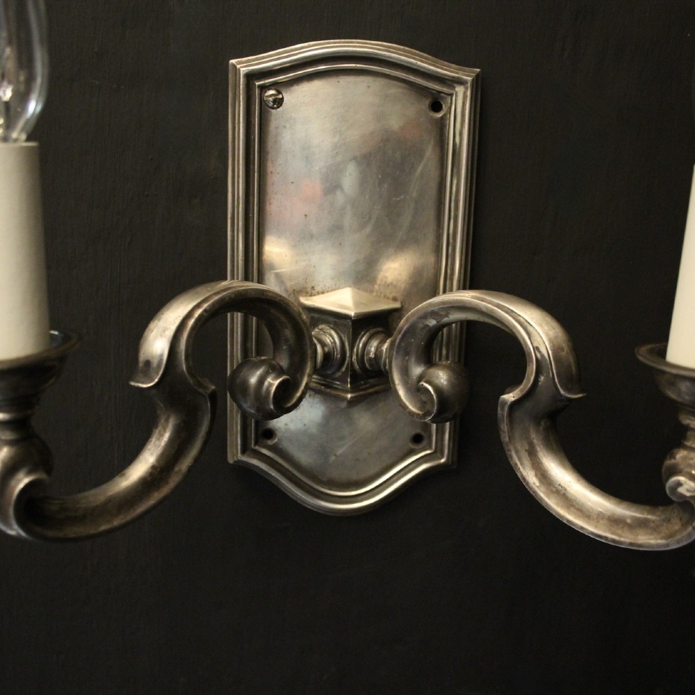 A pair of antique silver-plated twin-branch wall sconces featuring scrolling arms and moulded backplates against a dark background. View III