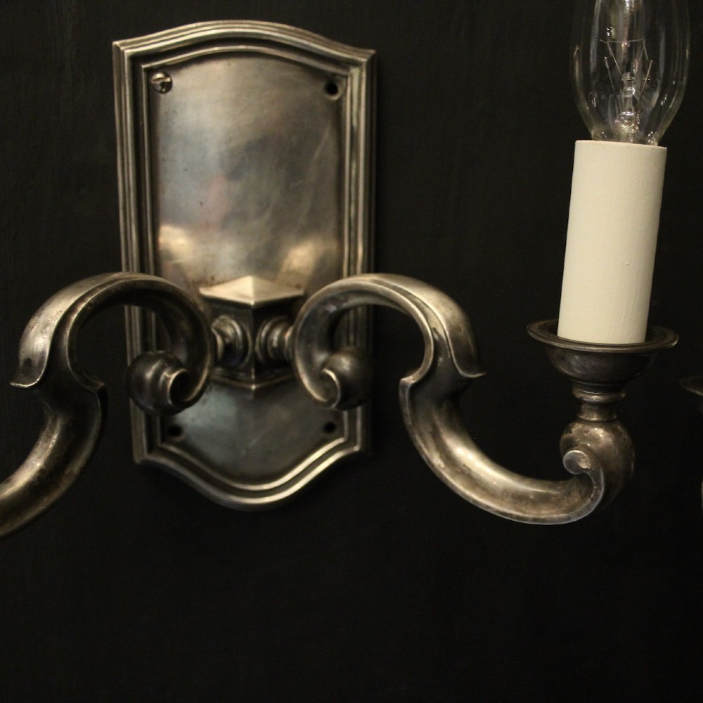 A pair of antique silver-plated twin-branch wall sconces featuring scrolling arms and moulded backplates against a dark background. View V