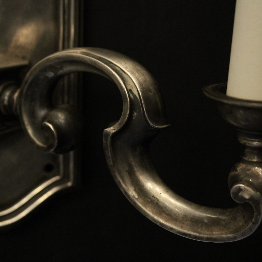 A pair of antique silver-plated twin-branch wall sconces featuring scrolling arms and moulded backplates against a dark background. View IX