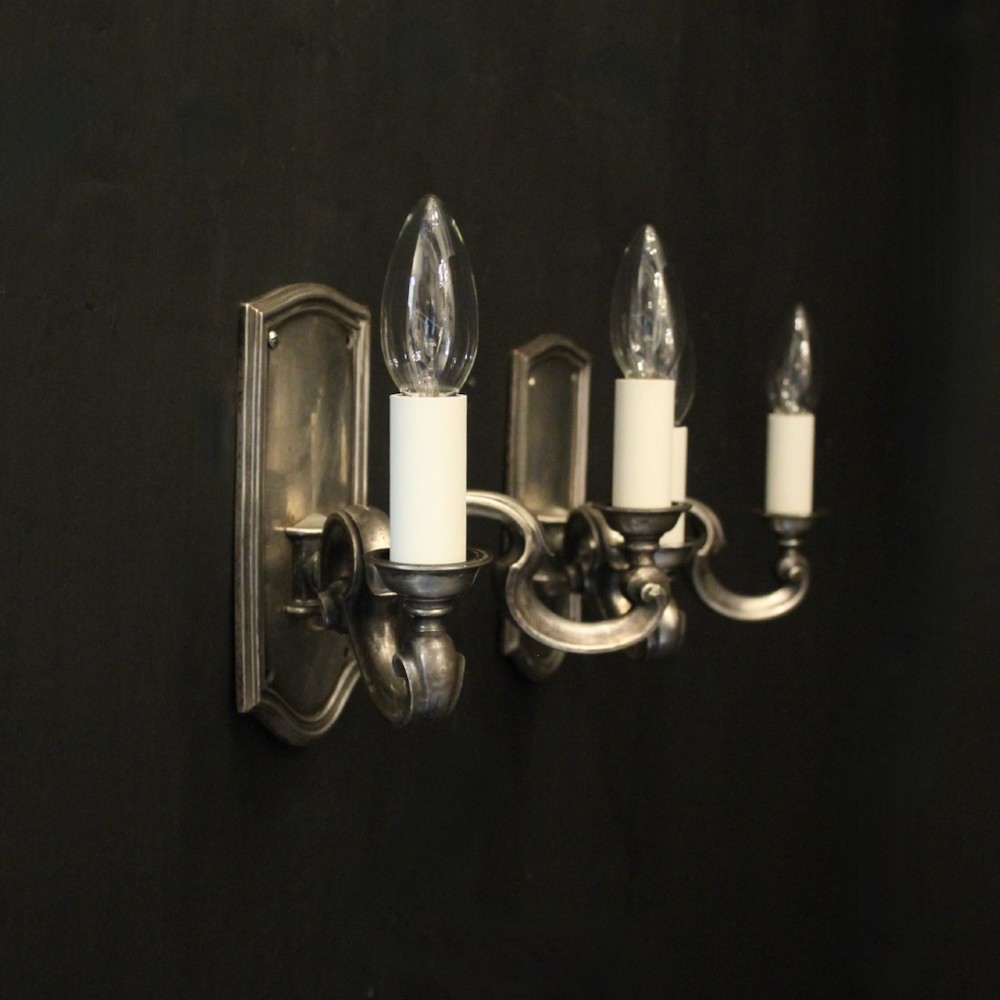 A pair of antique silver-plated twin-branch wall sconces featuring scrolling arms and moulded backplates against a dark background. View X