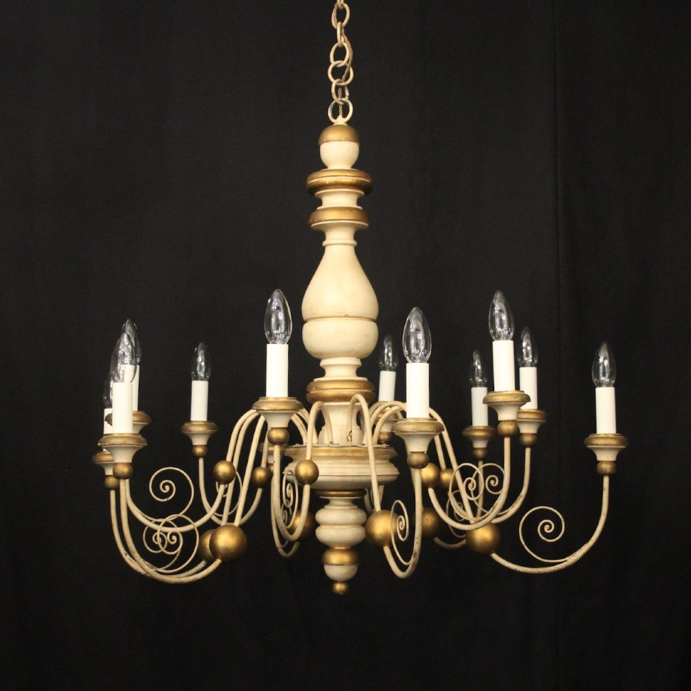 A twelve-light Florentine style ceiling chandelier featuring a cream and gilded turned wooden central column with wrought scrollwork branch arms and candle sleeves.