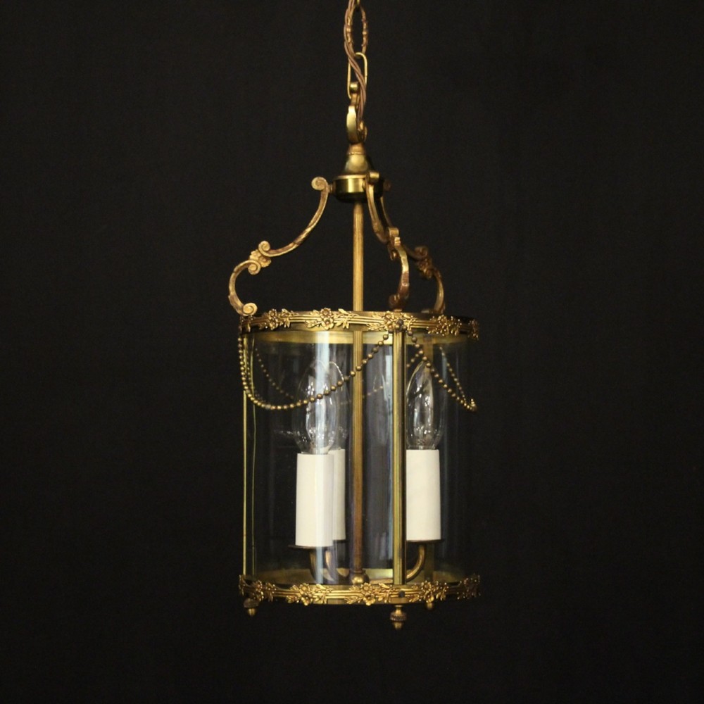 A cylindrical French gilded brass hall lantern featuring three central candle light fittings, curved top scrolls, floral garlands, and clear glass panes.