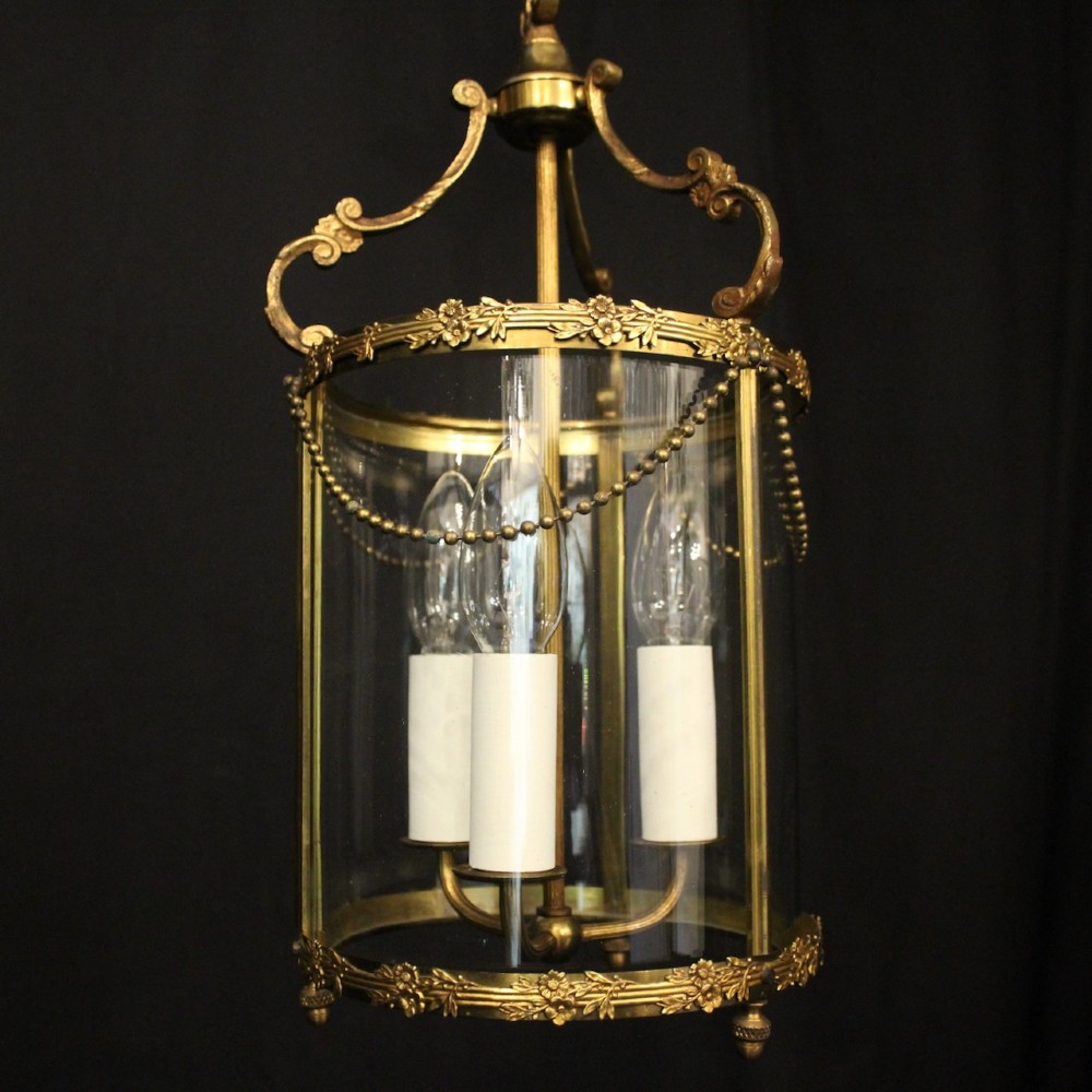 A cylindrical French gilded brass hall lantern featuring three central candle light fittings, curved top scrolls, floral garlands, and clear glass panes. View II
