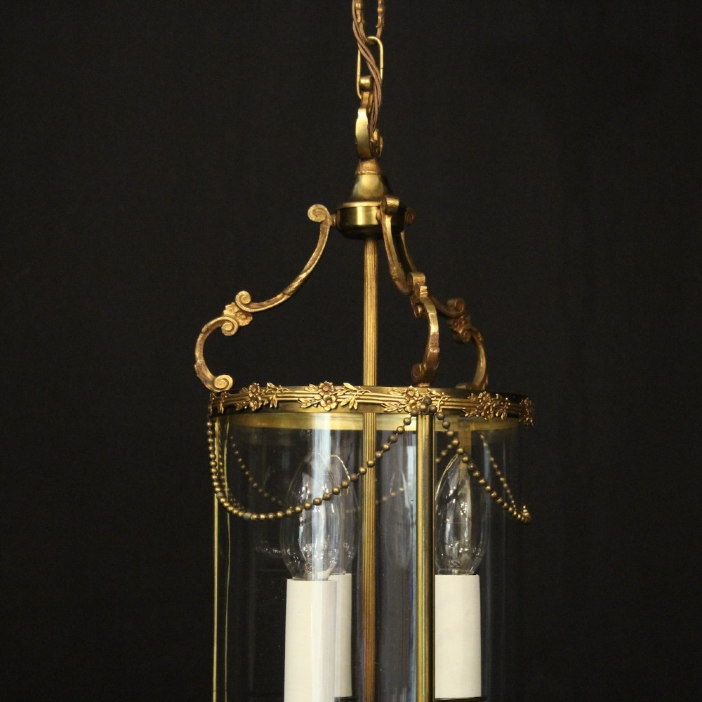 A cylindrical French gilded brass hall lantern featuring three central candle light fittings, curved top scrolls, floral garlands, and clear glass panes. View III