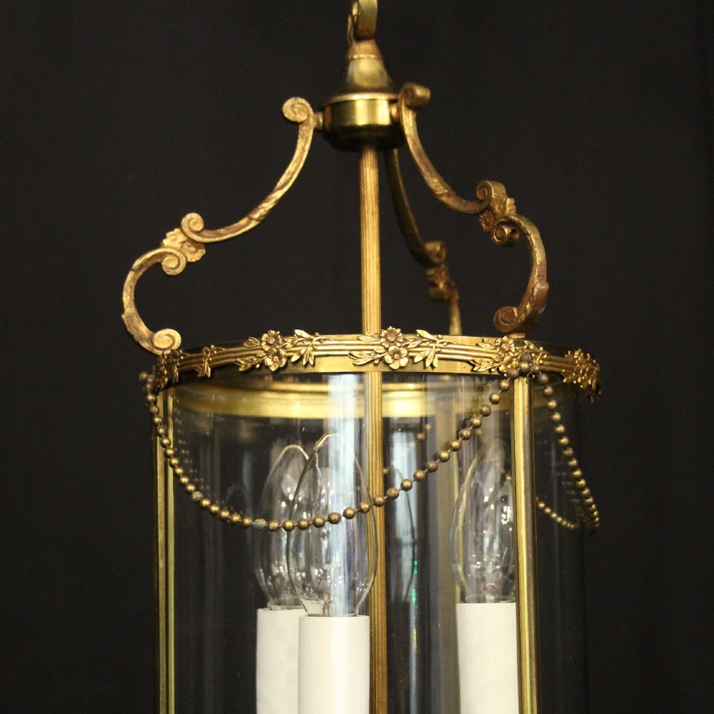 A cylindrical French gilded brass hall lantern featuring three central candle light fittings, curved top scrolls, floral garlands, and clear glass panes. View IV