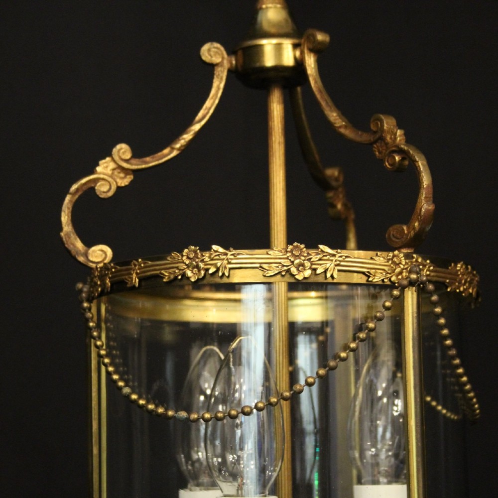 A cylindrical French gilded brass hall lantern featuring three central candle light fittings, curved top scrolls, floral garlands, and clear glass panes. View V