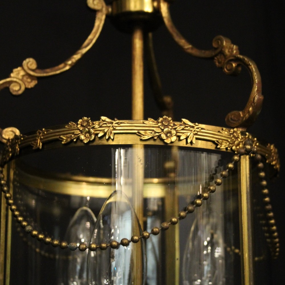 A cylindrical French gilded brass hall lantern featuring three central candle light fittings, curved top scrolls, floral garlands, and clear glass panes. View VI