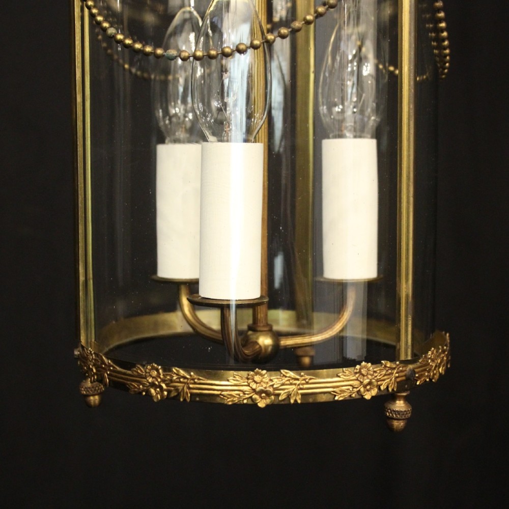 A cylindrical French gilded brass hall lantern featuring three central candle light fittings, curved top scrolls, floral garlands, and clear glass panes. View VII