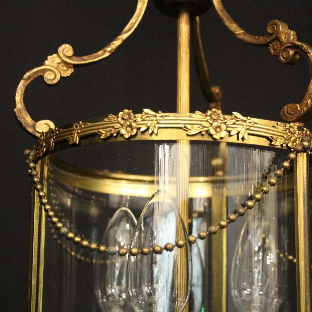 A cylindrical French gilded brass hall lantern featuring three central candle light fittings, curved top scrolls, floral garlands, and clear glass panes. View VIII