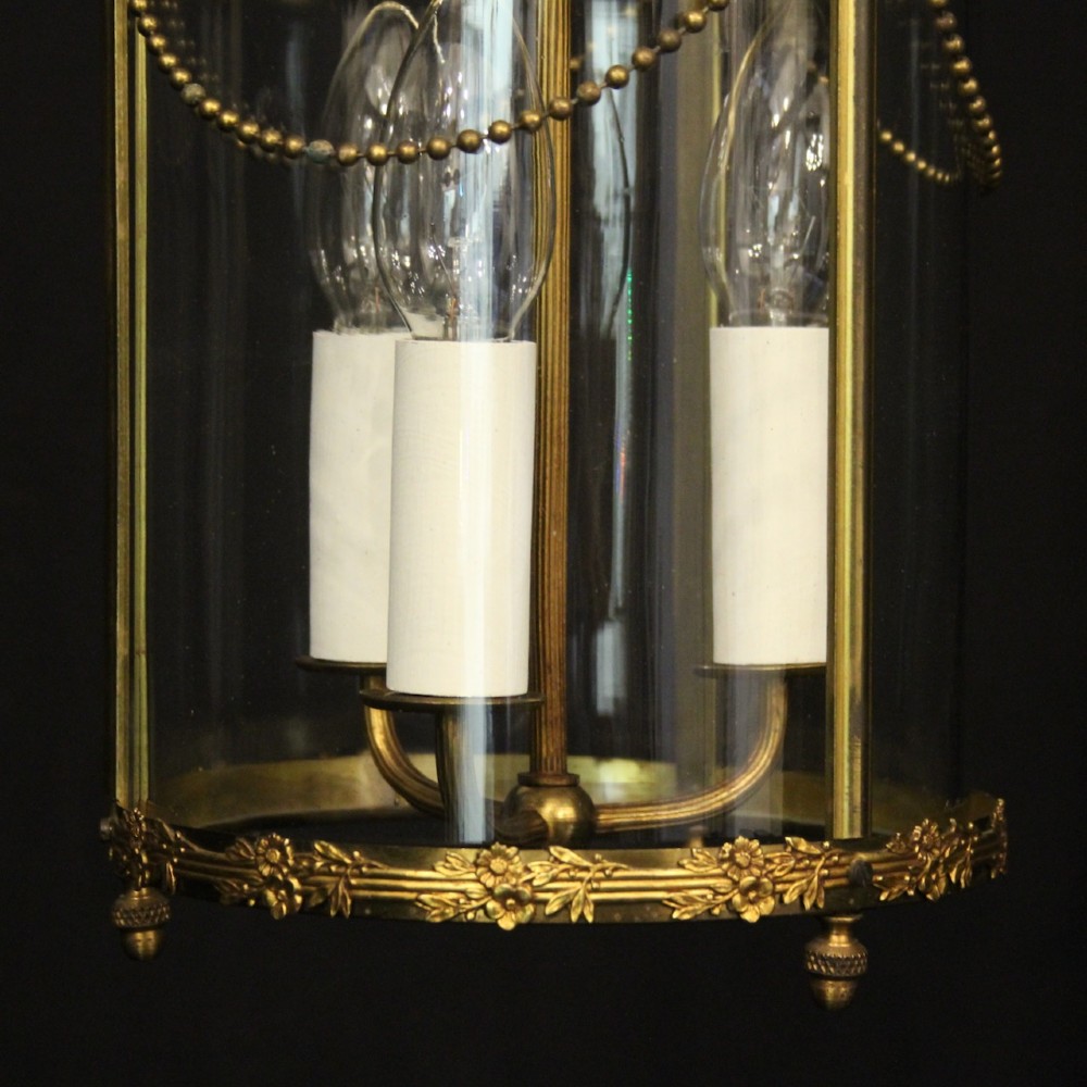 A cylindrical French gilded brass hall lantern featuring three central candle light fittings, curved top scrolls, floral garlands, and clear glass panes. View IX