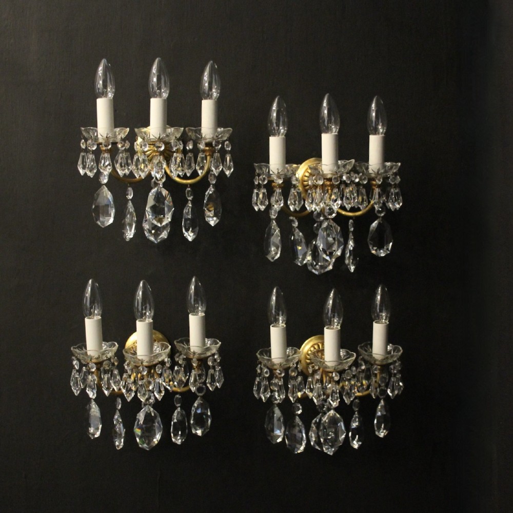 A set of four French triple-arm wall lights with gilt brass frames, candle sleeves, glass drip pans, and clear faceted crystal drops.