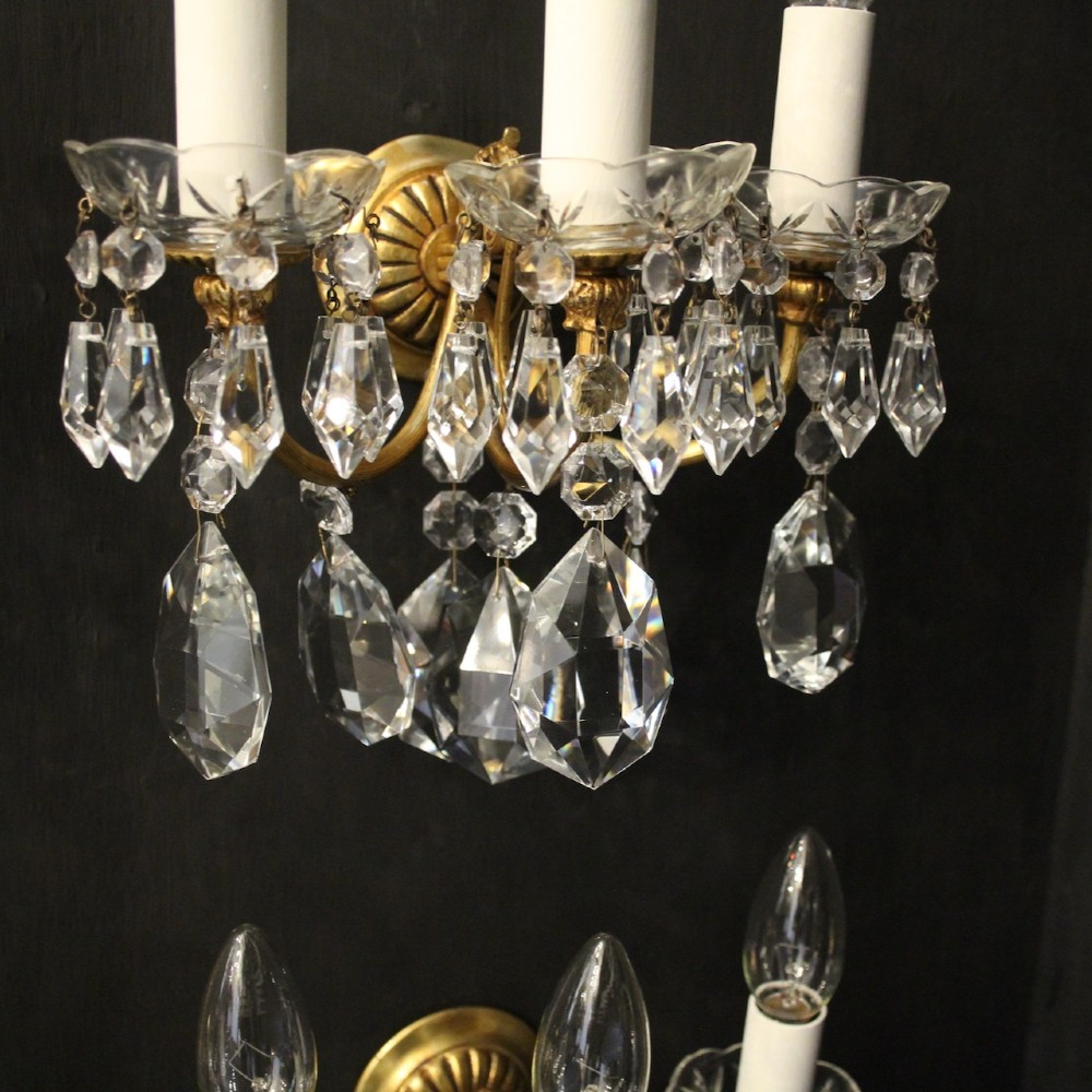 A set of four French triple-arm wall lights with gilt brass frames, candle sleeves, glass drip pans, and clear faceted crystal drops. View VII