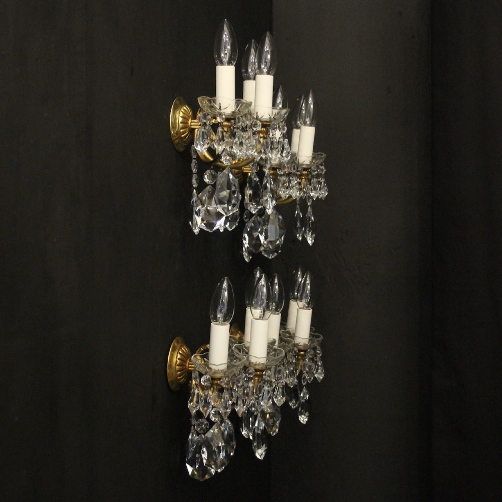 A set of four French triple-arm wall lights with gilt brass frames, candle sleeves, glass drip pans, and clear faceted crystal drops. View X