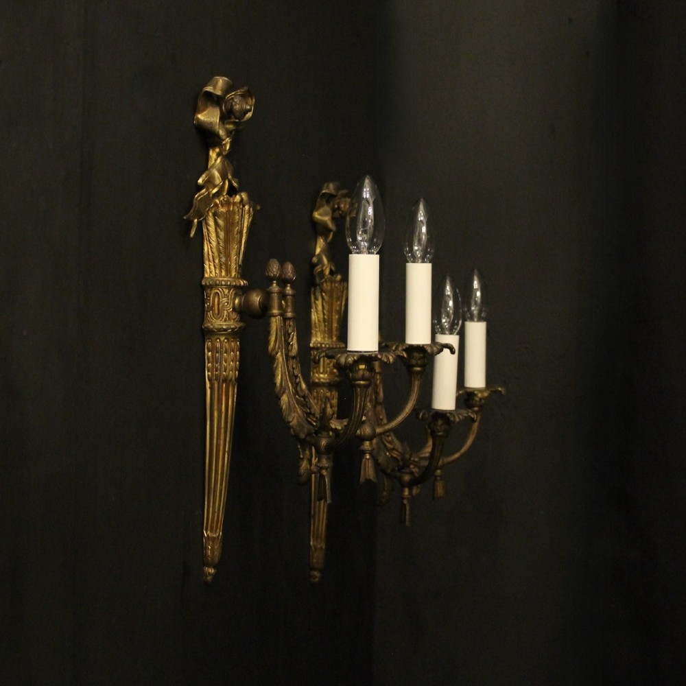A pair of French gilt bronze two-light wall sconces featuring ribbon bow crestings and fluted tapering backplates mounted on a dark background. View X