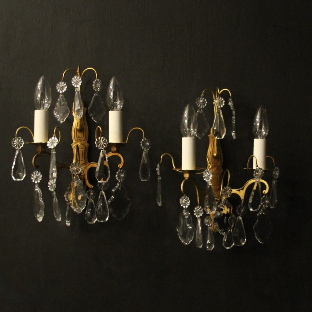 A pair of French gilded wall lights featuring twin candle-style bulb sockets, scrolling metal arms, and hanging faceted crystal droplets.