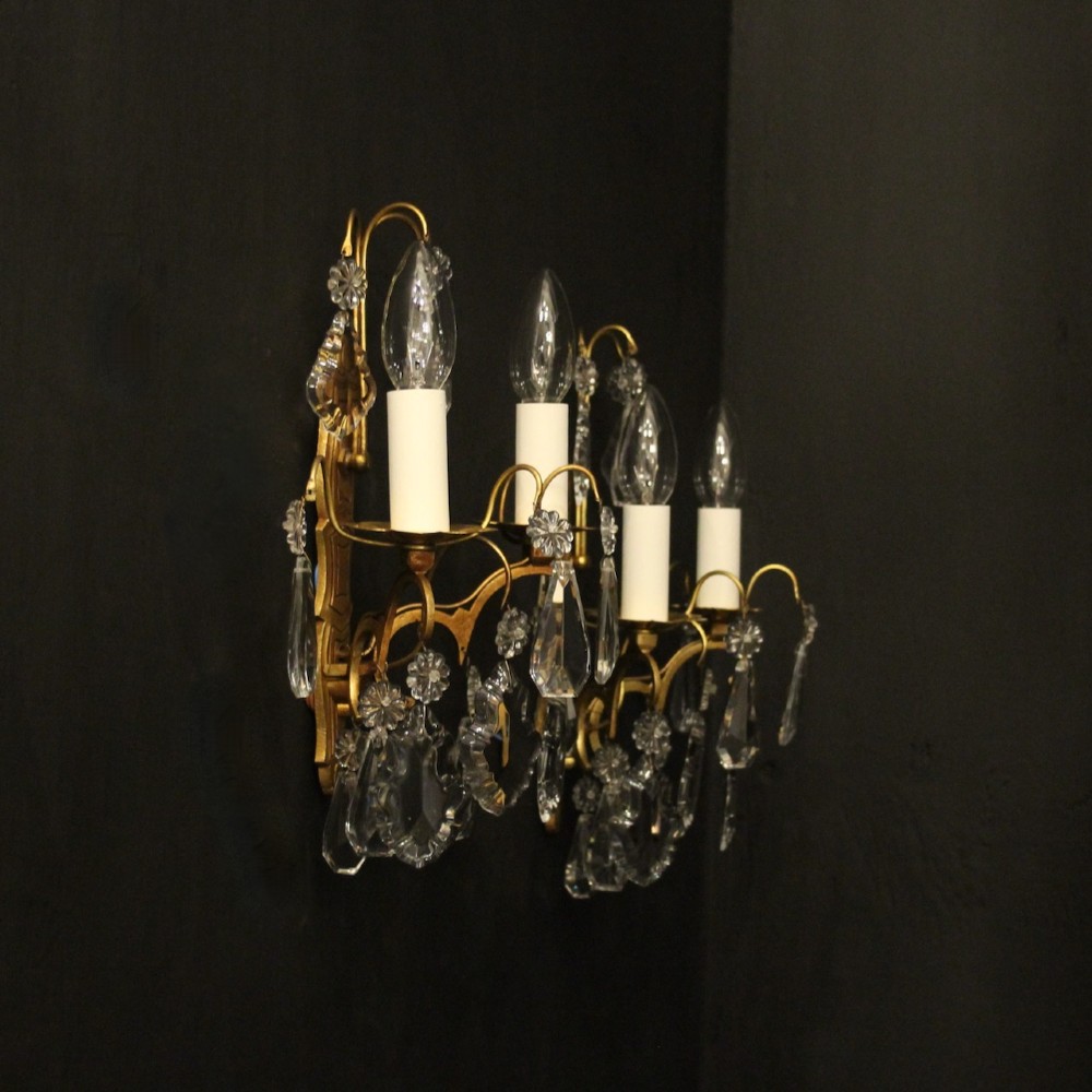 A pair of French gilded wall lights featuring twin candle-style bulb sockets, scrolling metal arms, and hanging faceted crystal droplets. View X