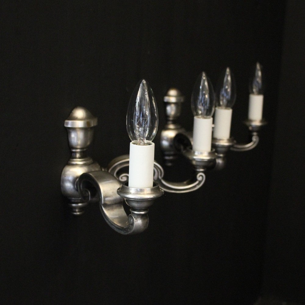 A pair of antique silver-plated twin-arm wall lights with scrolled branches, turned central urn backplates, and faux candle sleeves. View X