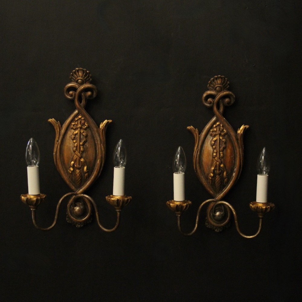 A pair of Florentine giltwood wall lights with twin curving arms, carved bow motifs, and faux candle bulb holders against a dark backdrop.