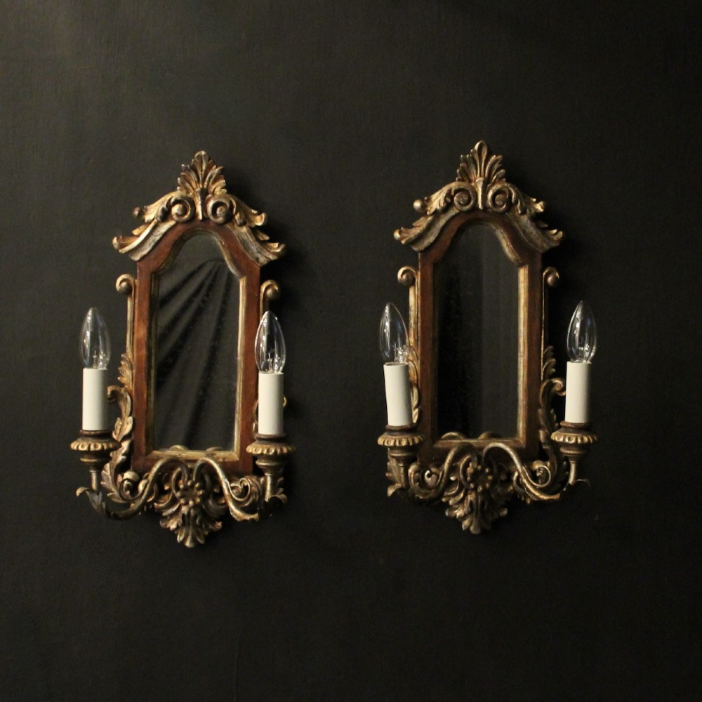 A pair of carved Florentine giltwood wall girandoles featuring arched mirror backplates, acanthus cresting, and twin light arms.
