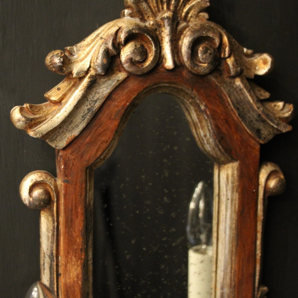 A pair of carved Florentine giltwood wall girandoles featuring arched mirror backplates, acanthus cresting, and twin light arms. View V