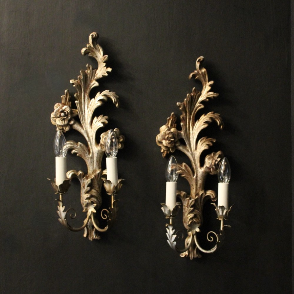 A pair of carved Florentine silver giltwood twin-arm wall sconces featuring acanthus leaf motifs and candle-style bulbs.