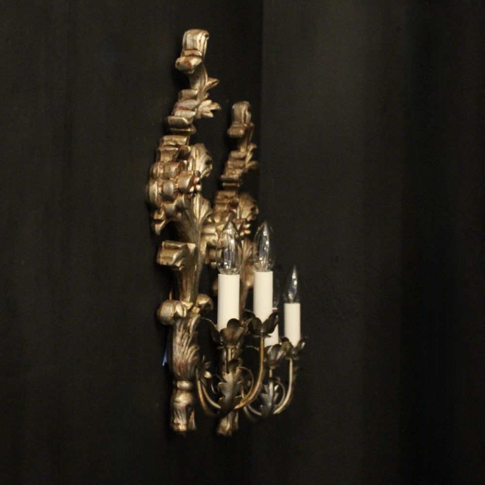 A pair of carved Florentine silver giltwood twin-arm wall sconces featuring acanthus leaf motifs and candle-style bulbs. View X