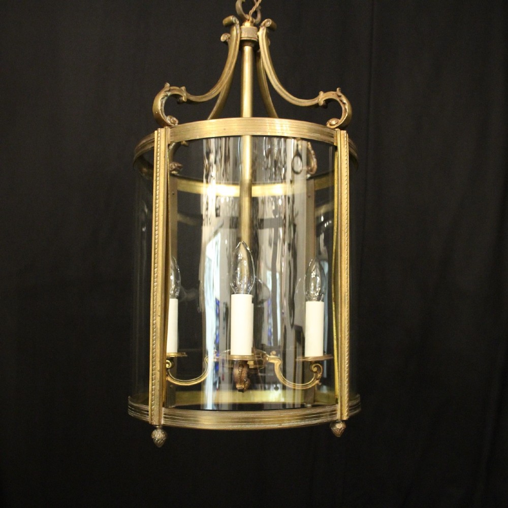A cylindrical gilded bronze hall lantern with curved clear glass panels and four internal candle bulb fittings hanging from a chain. View II
