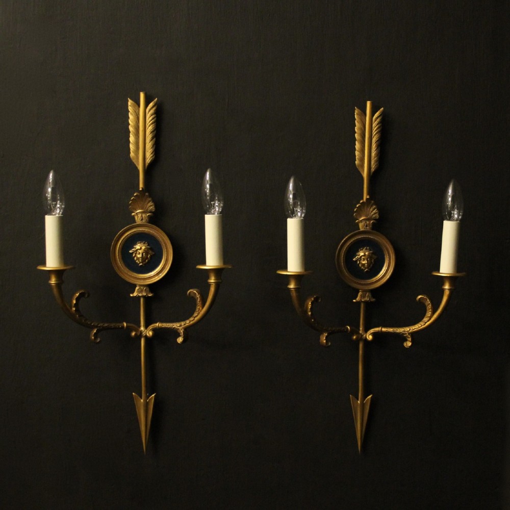 A pair of French gilt bronze twin-arm wall sconces featuring arrow-shaped backplates and central circular medallions with classical masks.