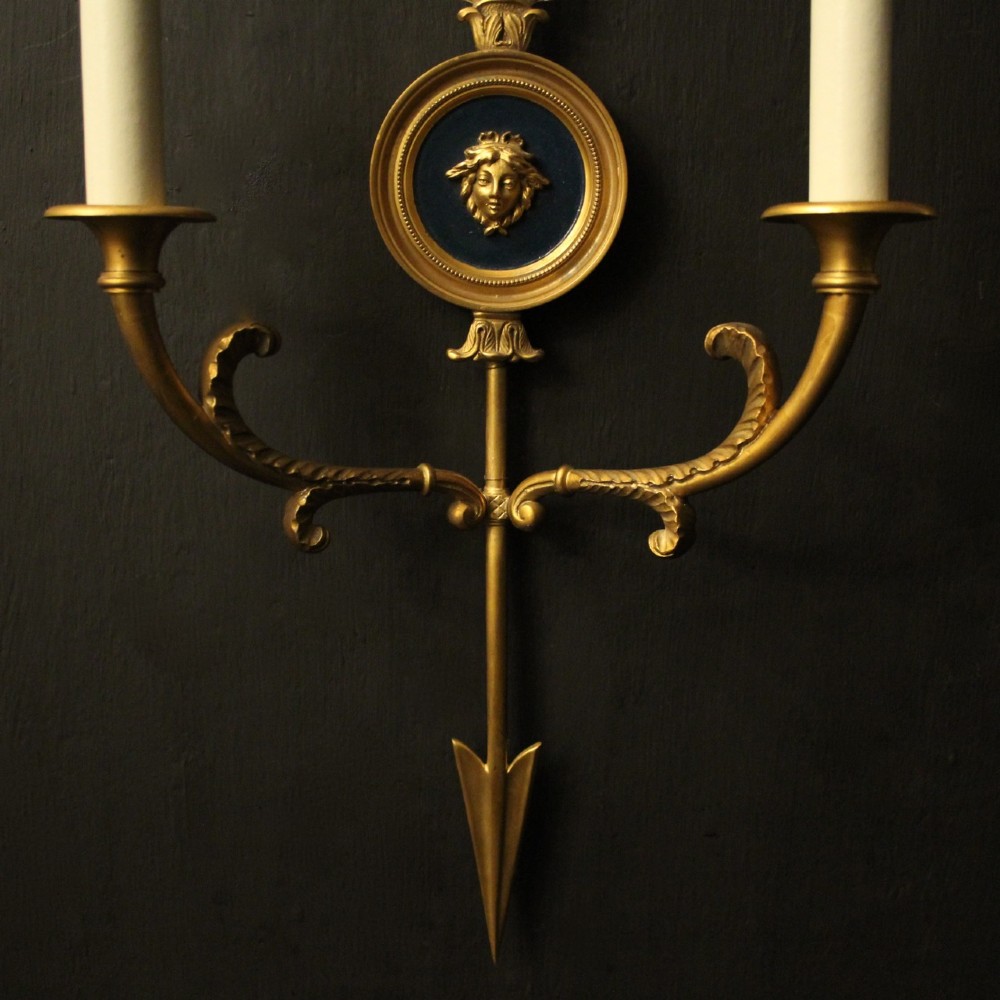 A pair of French gilt bronze twin-arm wall sconces featuring arrow-shaped backplates and central circular medallions with classical masks. View VI
