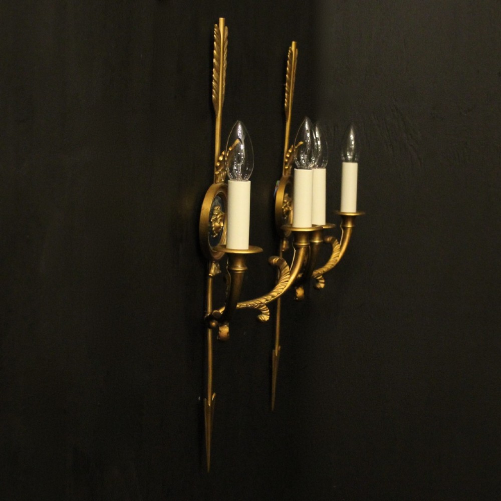 A pair of French gilt bronze twin-arm wall sconces featuring arrow-shaped backplates and central circular medallions with classical masks. View X