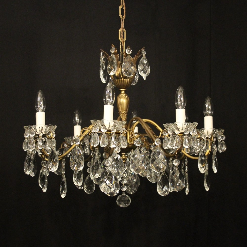 An eight-arm gilded metal chandelier suspended from a chain, decorated with faceted glass droplets and glass candle-style light holders.