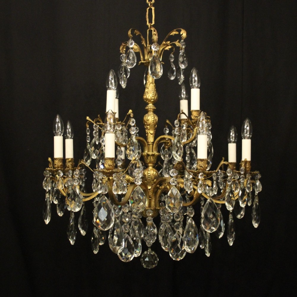 A French gilded bronze twelve-light chandelier featuring curved scrolling arms, cream candle-style light holders, and cascading faceted crystal drops.