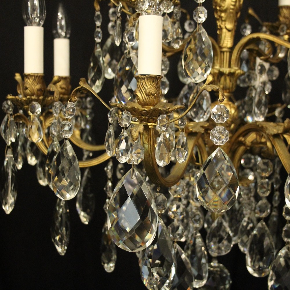 A French gilded bronze twelve-light chandelier featuring curved scrolling arms, cream candle-style light holders, and cascading faceted crystal drops. View VI