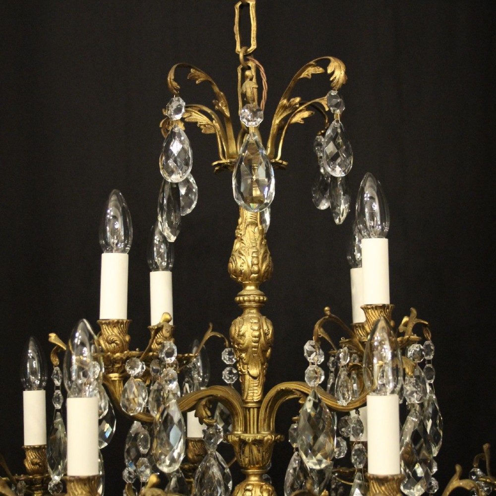 A French gilded bronze twelve-light chandelier featuring curved scrolling arms, cream candle-style light holders, and cascading faceted crystal drops. View IX
