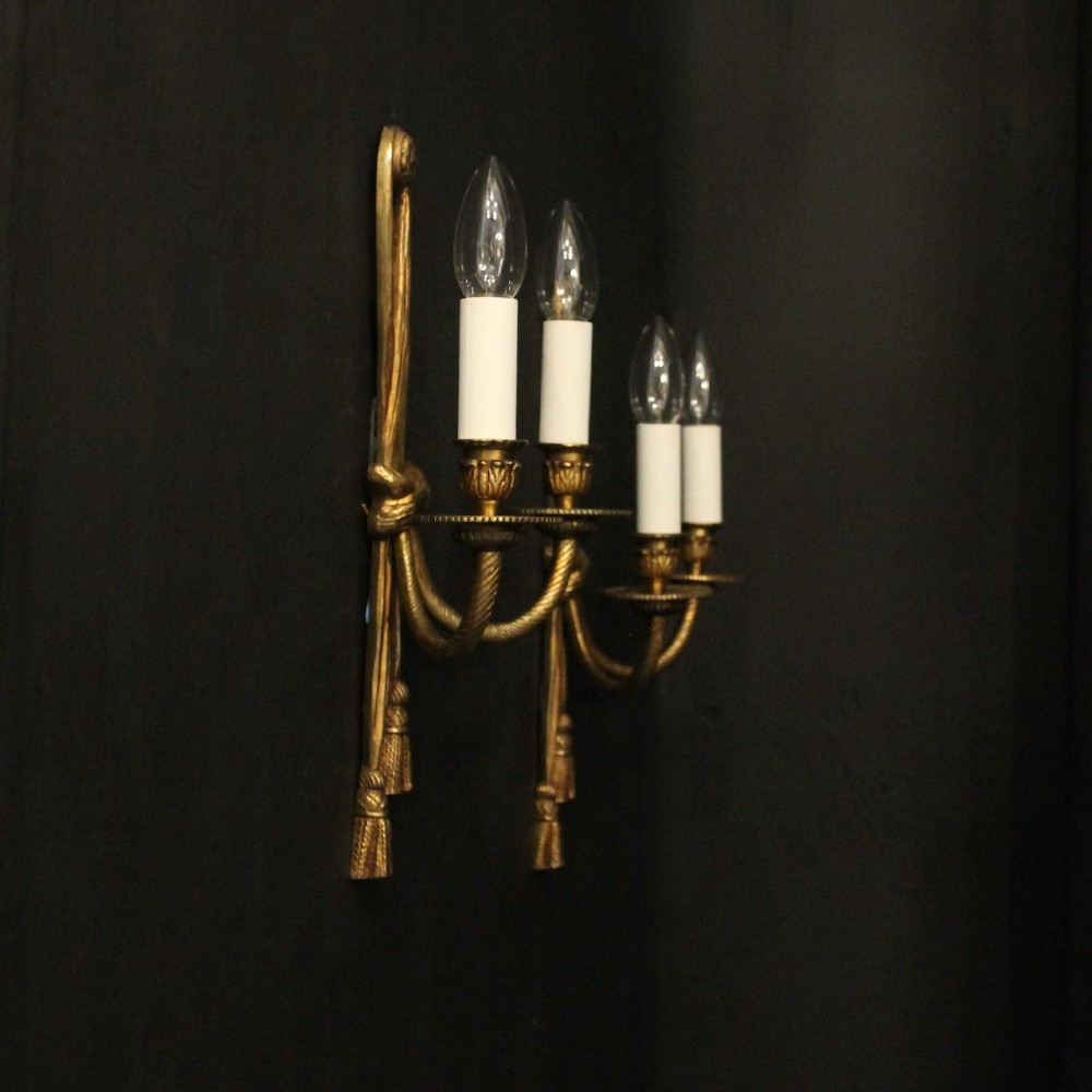 A pair of French gilded brass twin-arm wall sconces featuring a tied rope and tassel motif with candle-style bulb fittings. View X