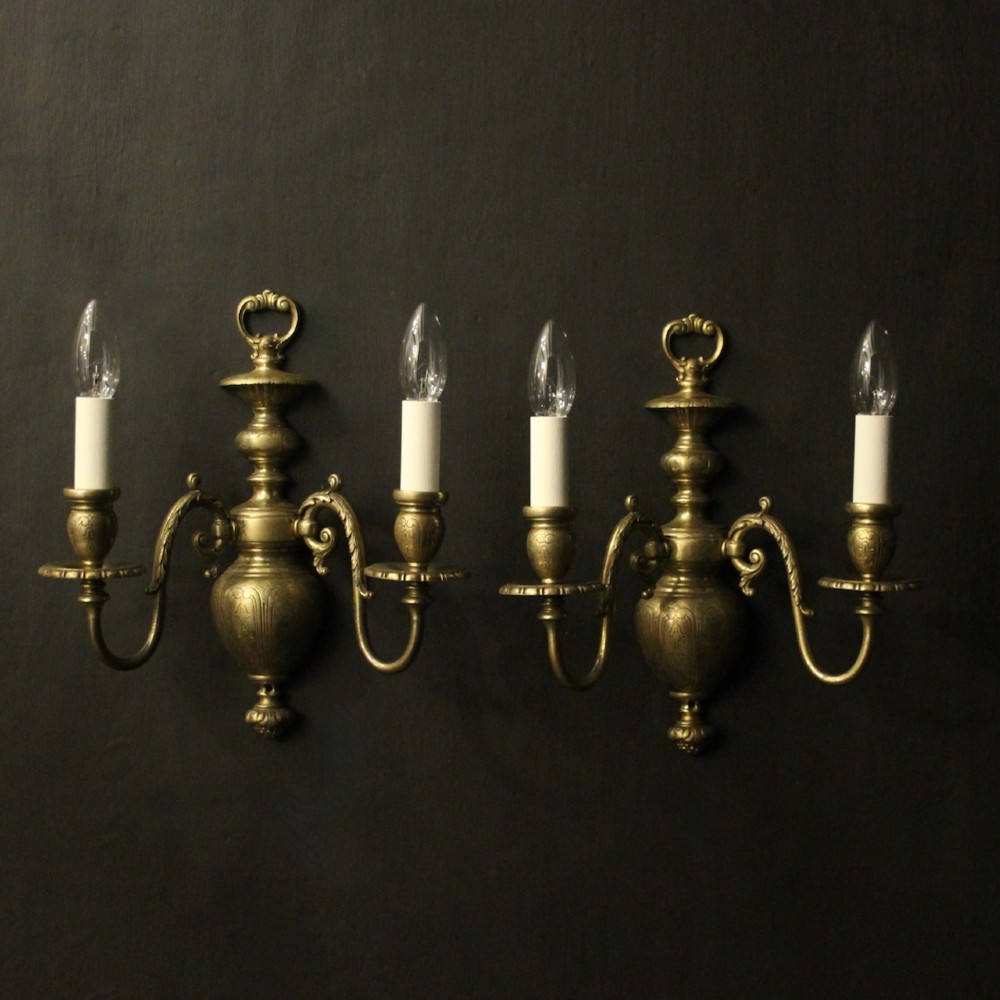 A pair of antique bronze twin-arm wall lights with urn-shaped central bodies and decorative scroll arms against a dark backdrop.