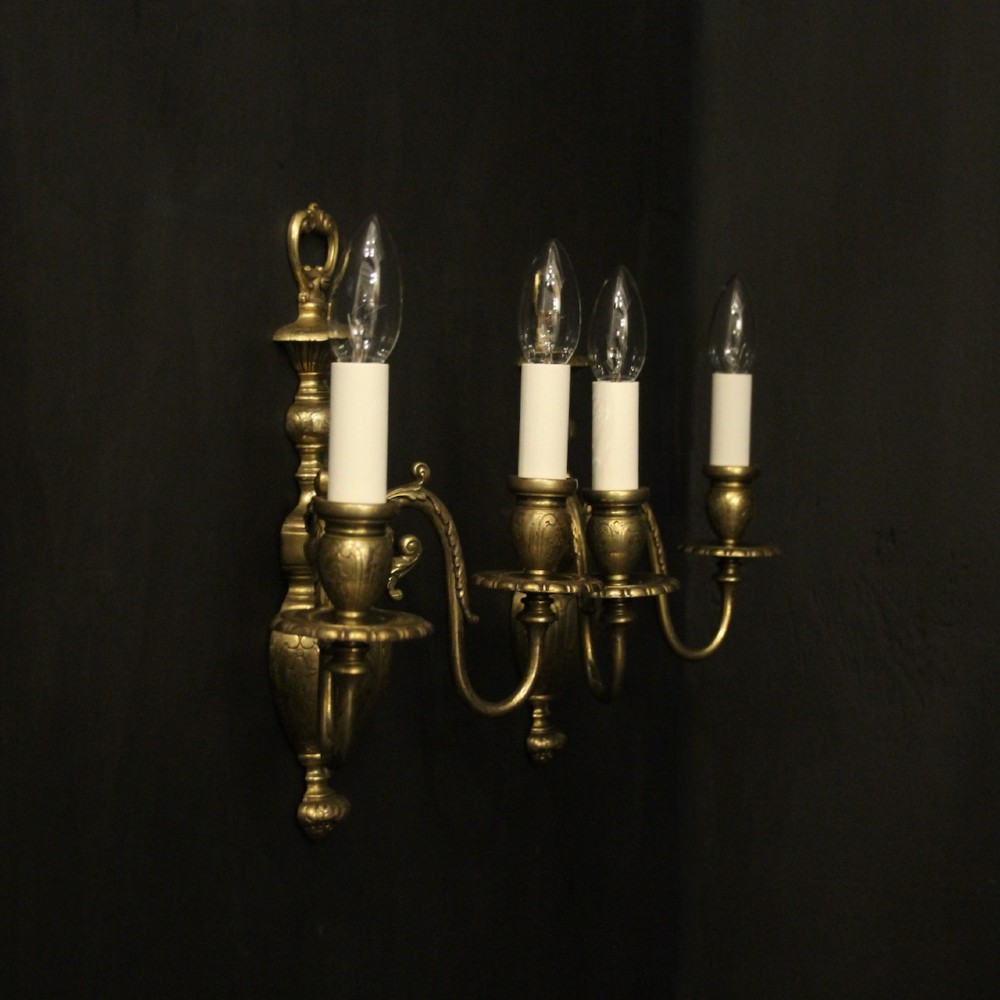 A pair of antique bronze twin-arm wall lights with urn-shaped central bodies and decorative scroll arms against a dark backdrop. View X