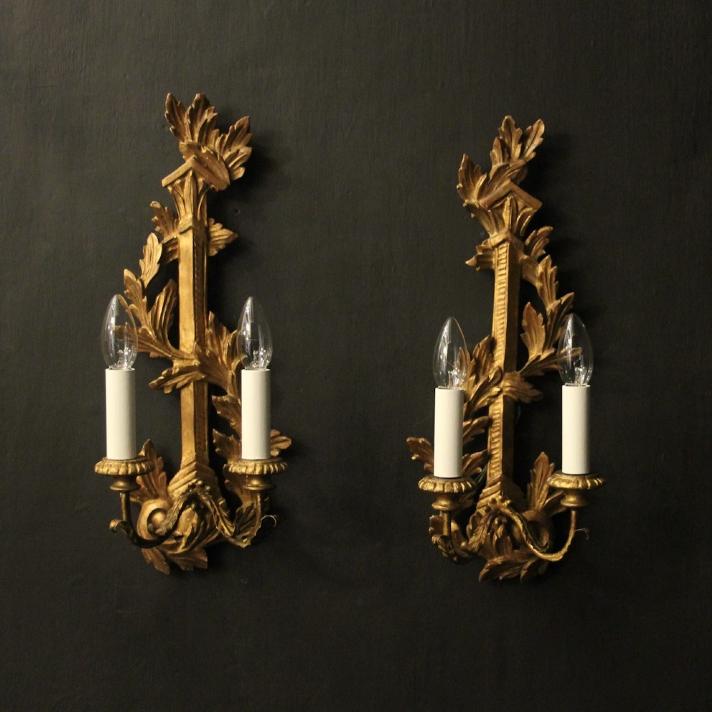 A pair of French carved giltwood two-arm wall sconces featuring leaf ornamentation and white candle sleeves with clear light bulbs.